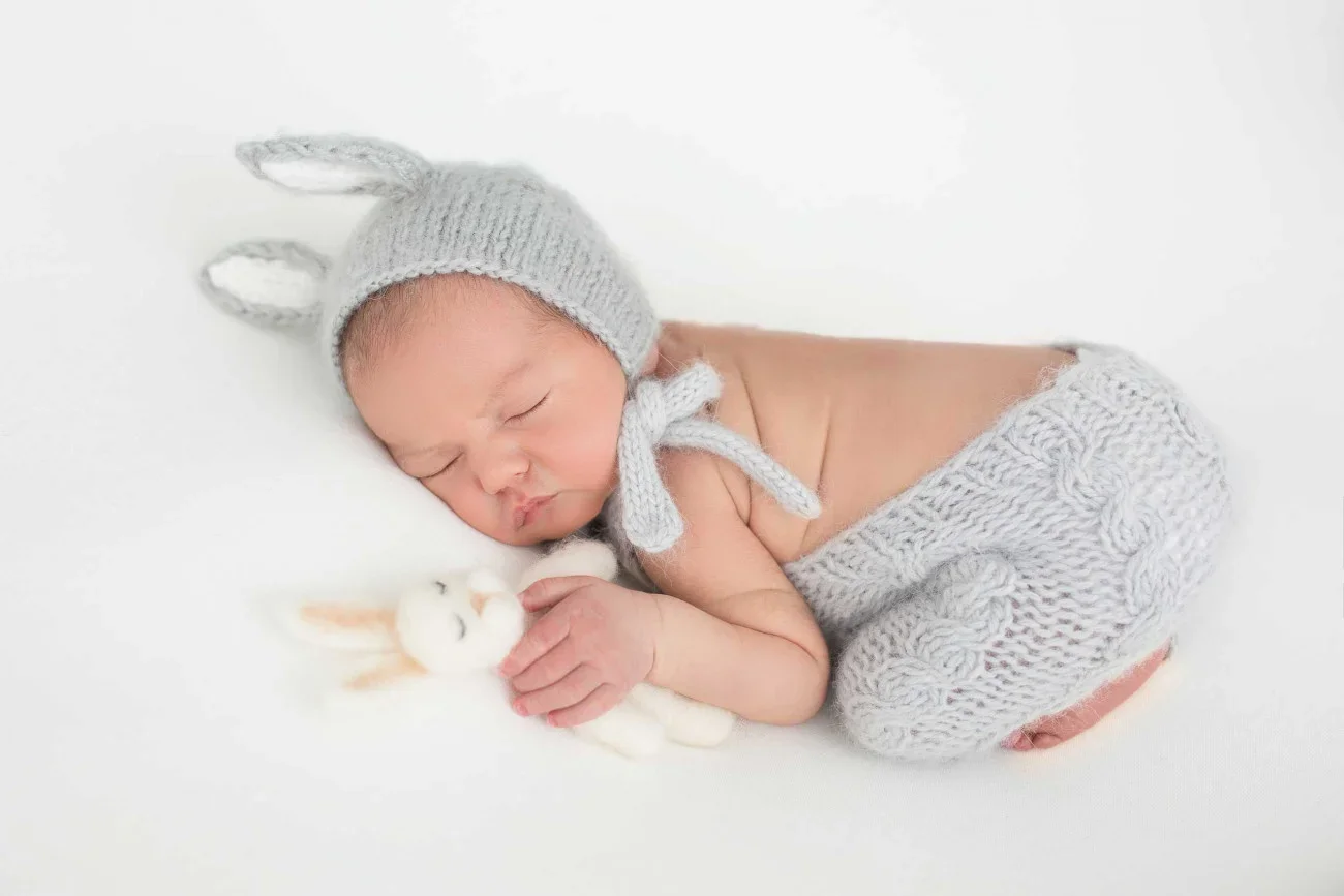 Newborn baby sleeping on white surface, wearing a knitted gray hat with bunny ears and matching pants, holding a small white plush bunny.