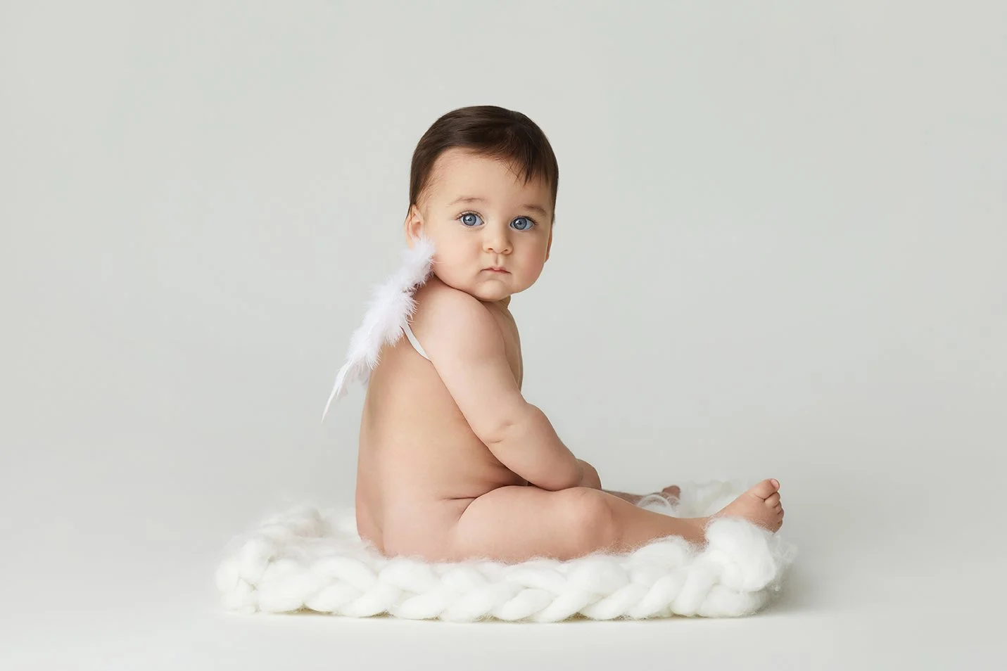 A baby with blue eyes sitting on a knitted woolen rug, wearing a white feathered angel wing accessory on one shoulder, against a plain light background.