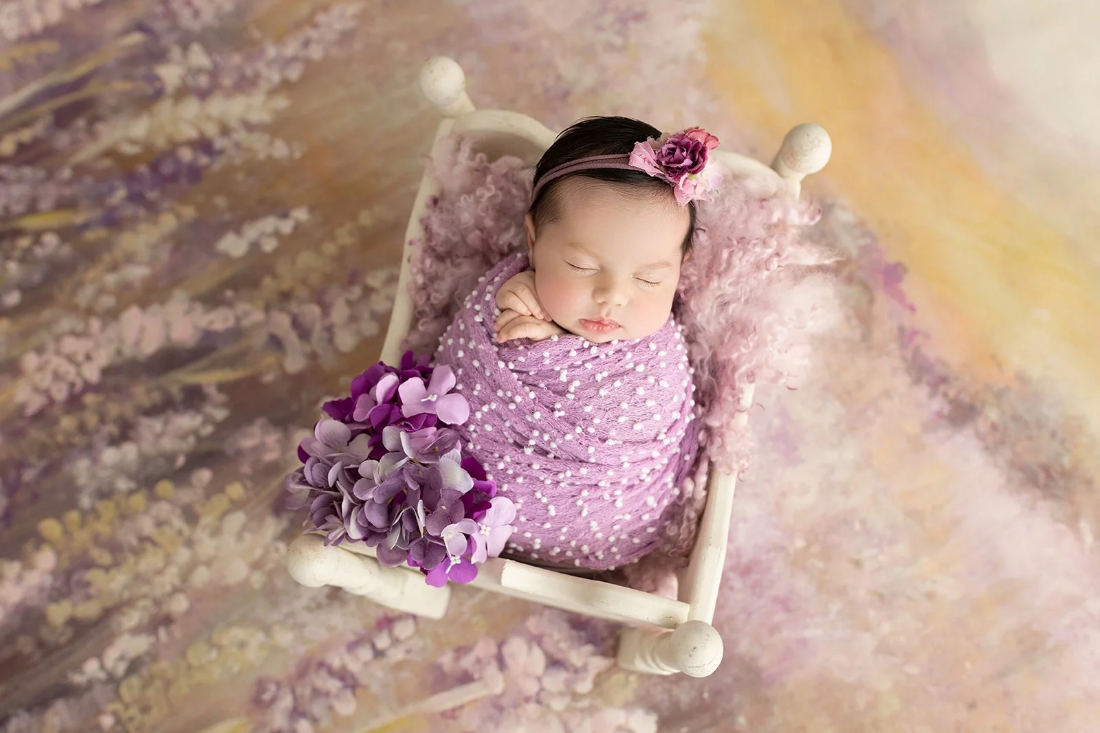 A sleeping baby girl wrapped in a pink blanket with white dots, adorned with a pink headband with flowers, and holding a bouquet of purple flowers, lying in a small white bed with a pink pillow and a pink furry blanket, on a soft, colorful background