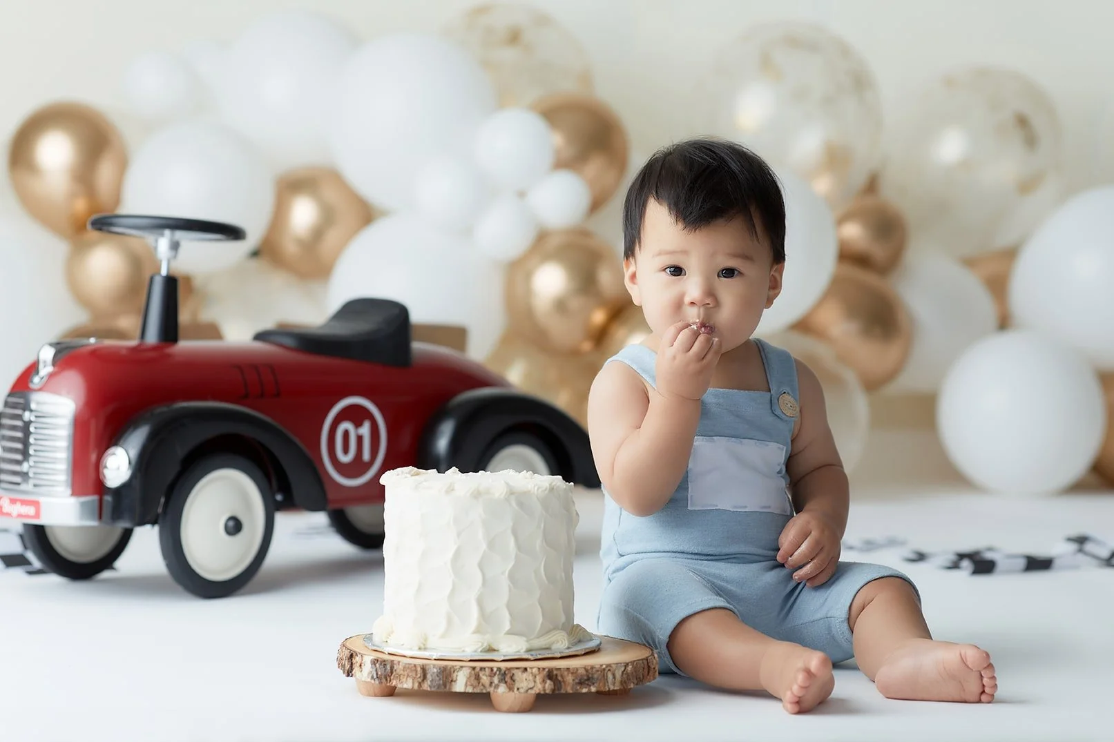 Young child sitting on the floor with a small white cake in front, surrounded by white and gold balloons, a red toy car, and a black and white checkered banner in the background.
