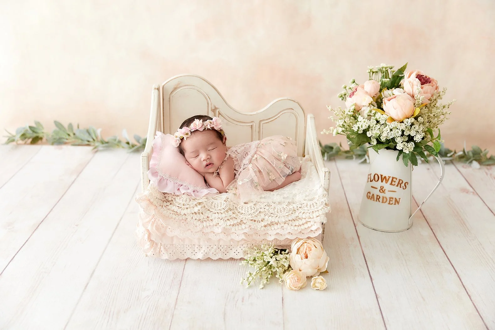 A sleeping baby girl lies on a lace pillow in a miniature white bed, wearing a pink lace dress and a flower crown, with a flower arrangement in a white pitcher and more flowers in front, on a white wooden floor with a soft pink background.