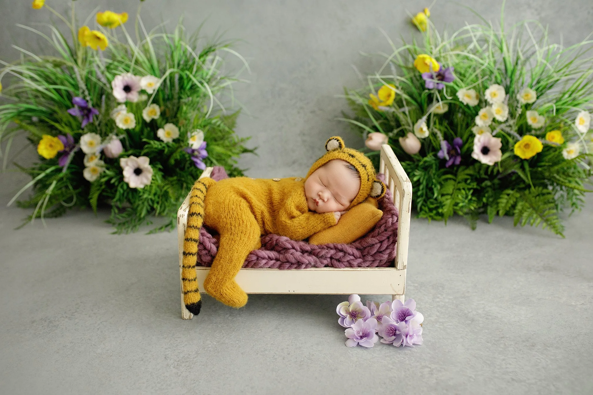 A sleeping baby dressed in a yellow tiger costume with a hood featuring tiger ears and tail, lying on a small bed with a pink blanket and an orange pillow, surrounded by purple and white flowers and green foliage.