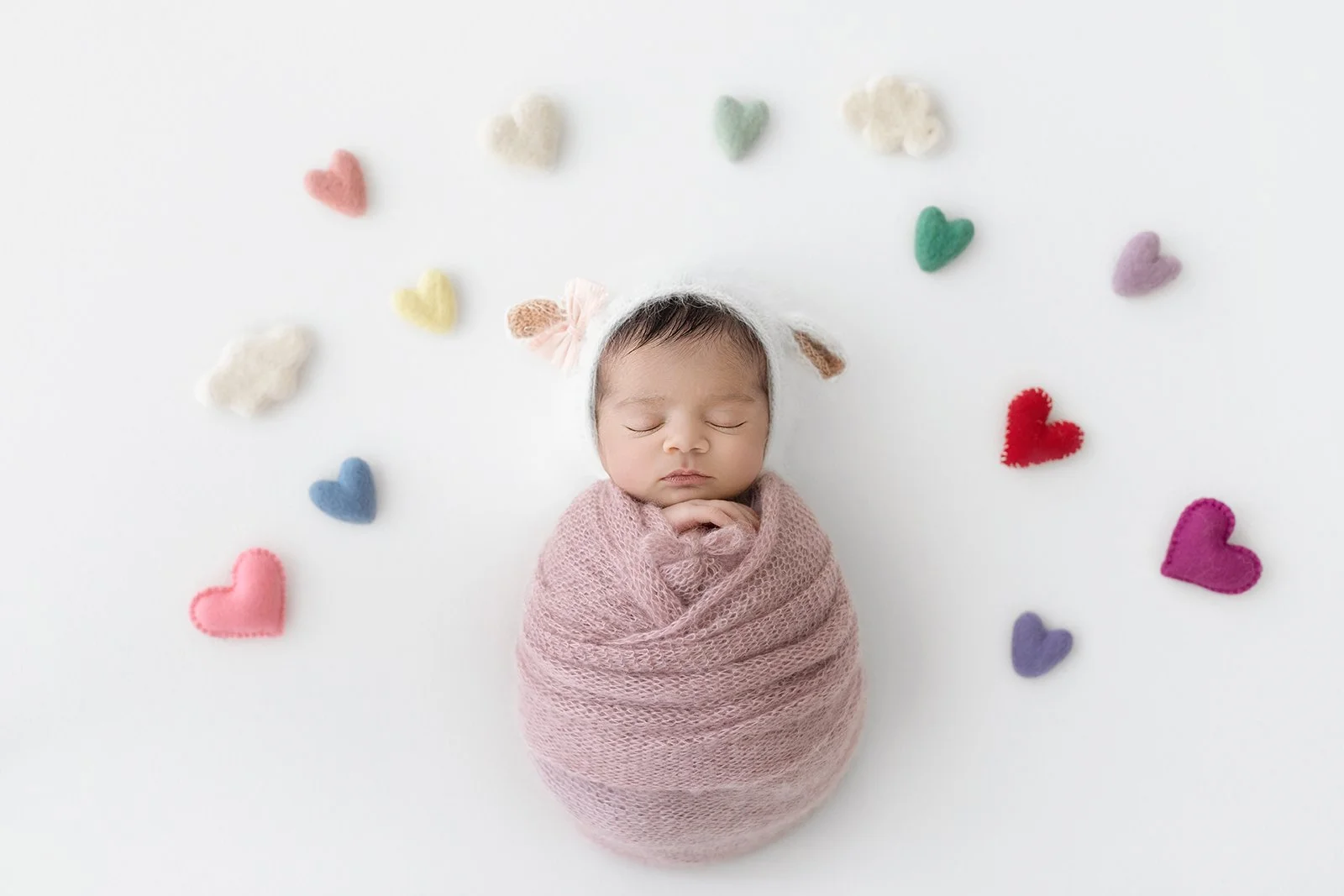 A sleeping baby wrapped in a pink blanket, wearing a white hat with bear ears, surrounded by small felt hearts and clouds in pastel colors on a white background.