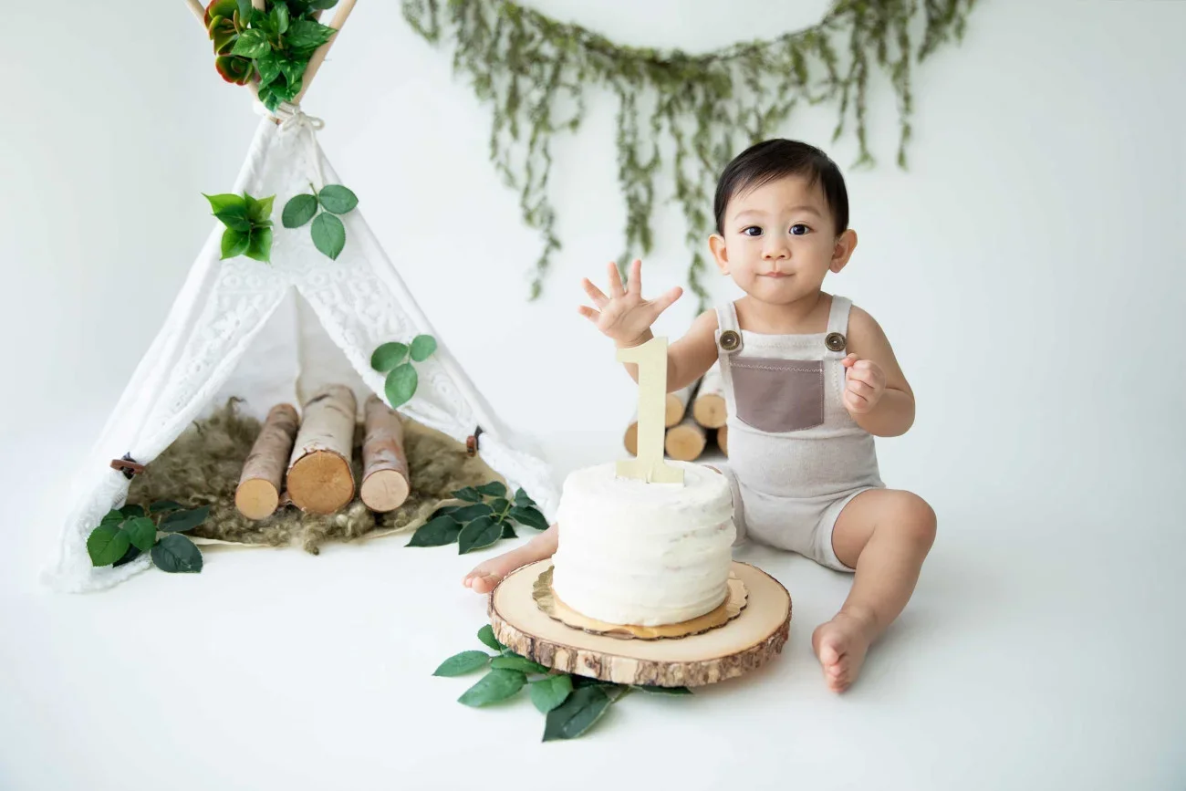 Baby celebrating first birthday with a cake and a wooden teepee tent in the background, decorated with green leaves and berries.