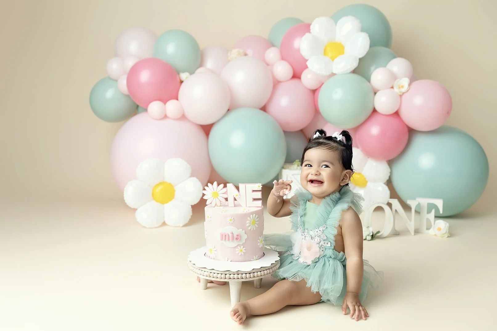 A young girl in a mint green dress celebrating her first birthday, sitting next to a pink birthday cake with the word 'ONE' on top, surrounded by pink, mint green, and white balloons, large paper flowers, and decorative letters spelling 'ONE' in the 