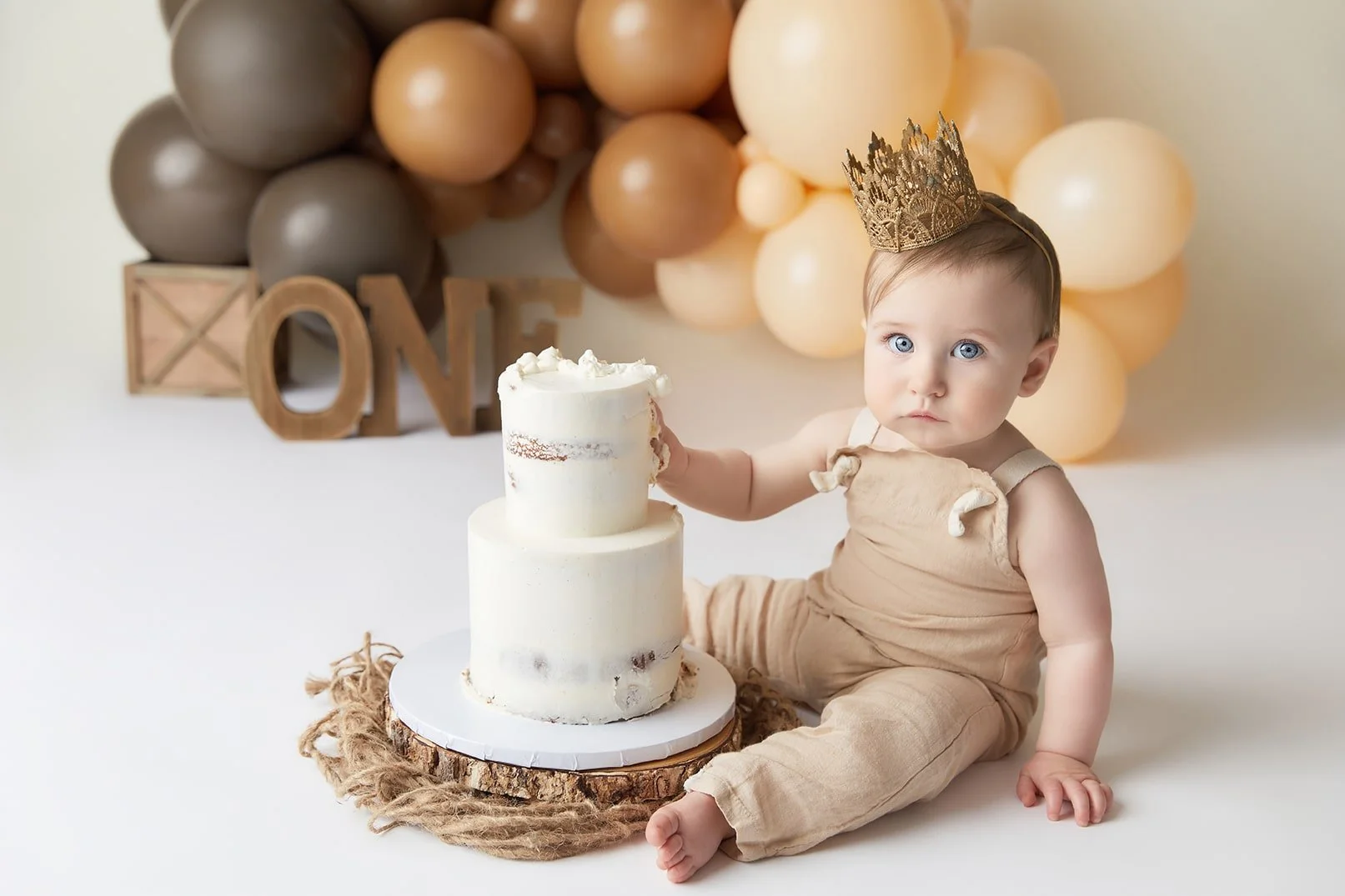 Baby sitting on the floor with a partially eaten birthday cake, wearing beige overalls and a gold lace crown, with balloons and wooden letters spelling 'LOVE' in the background.