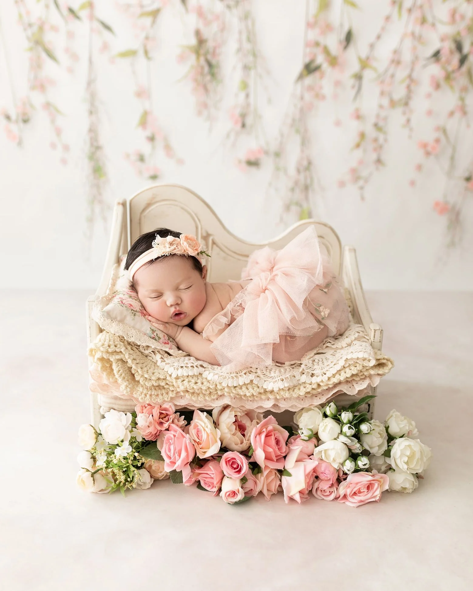 A sleeping baby girl lying on a small vintage white bed, adorned with pink and white flowers, and a pink tulle dress with a matching headband.