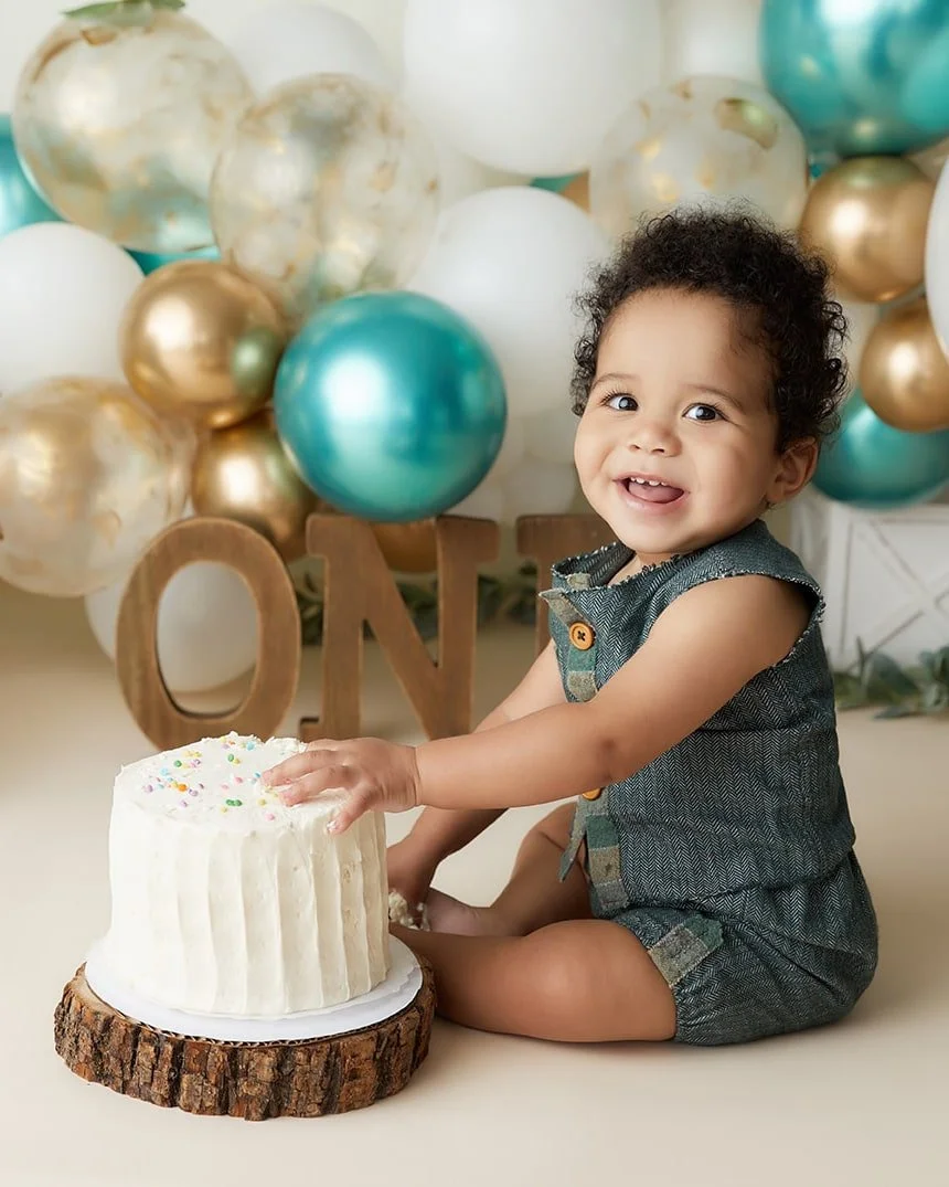 A smiling toddler with curly hair celebrating birthday with a small cake, surrounded by balloons in gold, white, and teal colors, with wooden letters spelling "ONE" in the background.