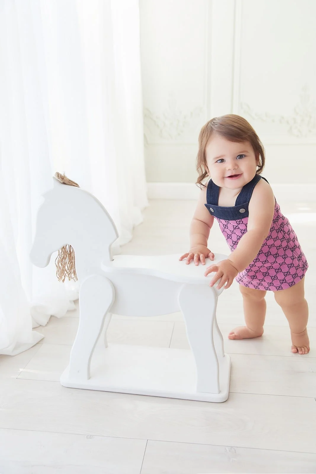 Smiling toddler girl in pink and purple dress with navy straps playing with a white wooden horse on a white floor in a bright room.