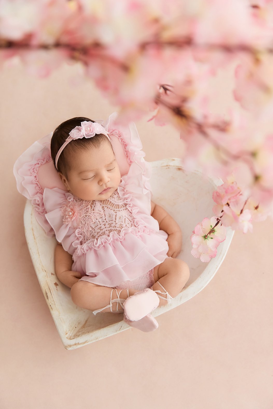 A sleeping baby girl in a pink dress and matching headband, lying in a small white wooden tub with pink flowers hanging above.