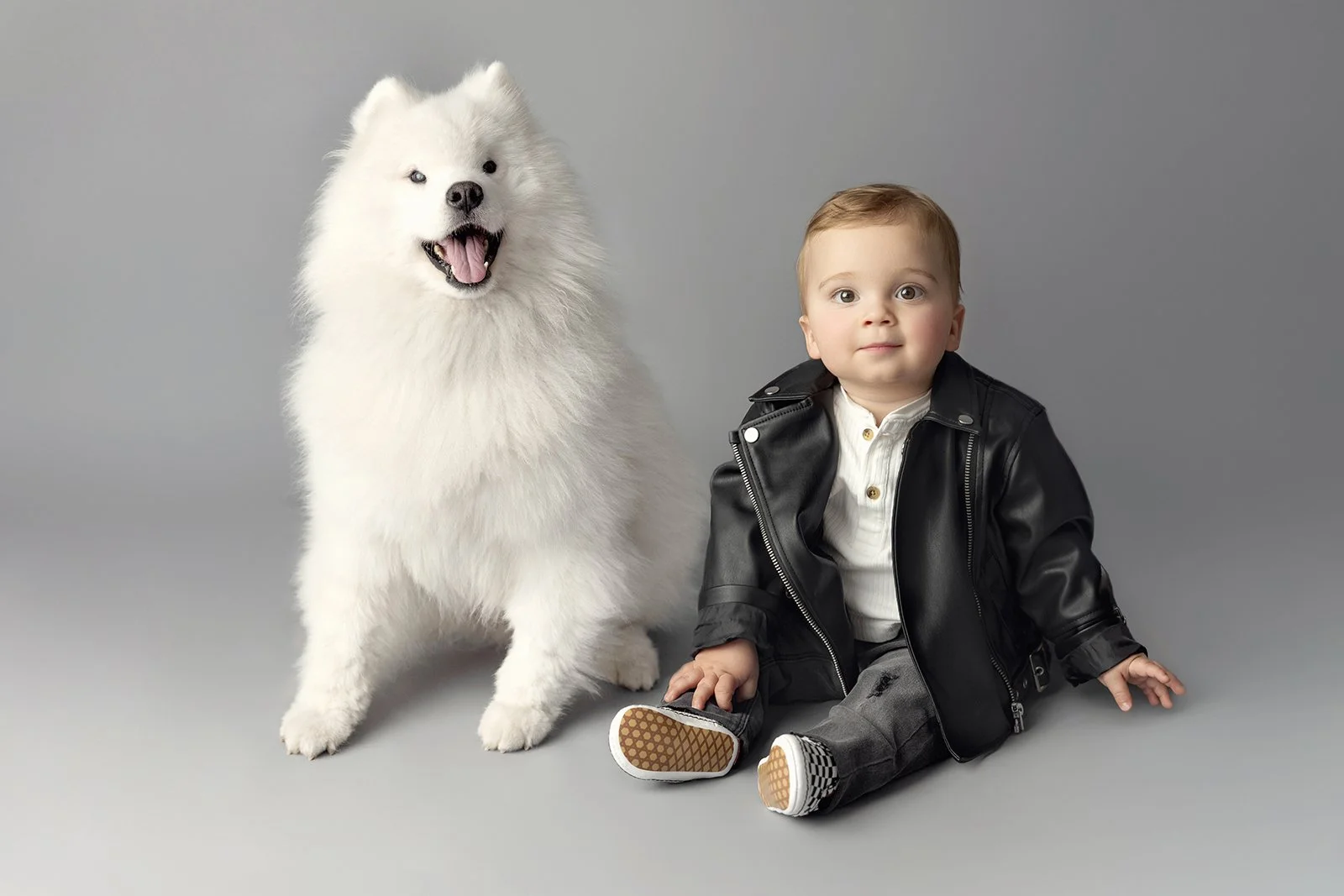A young boy sitting on the floor next to a large, fluffy white dog, both facing the camera against a plain gray background.