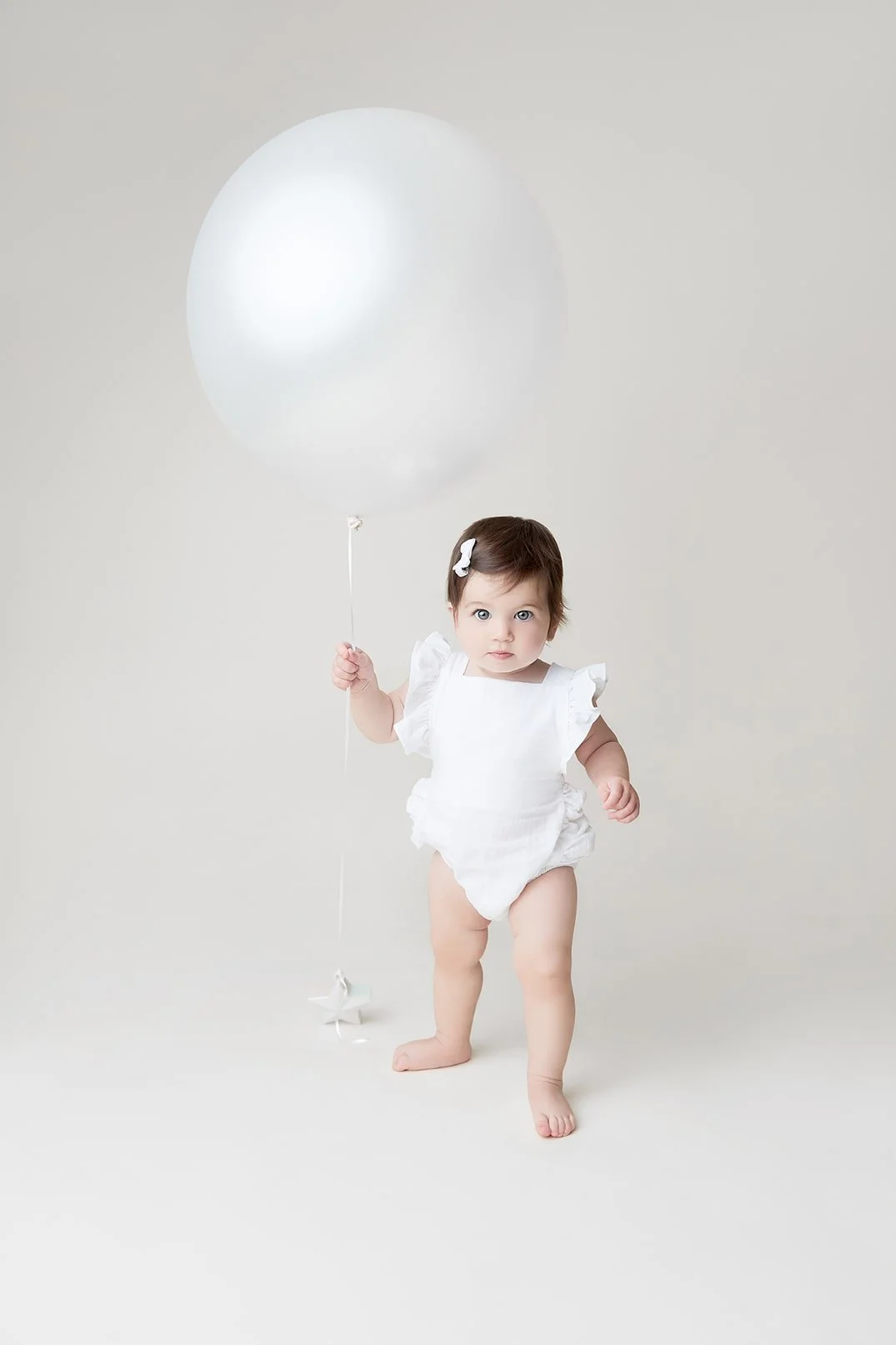 A baby girl with a white dress and hair clip holding a large white balloon in a minimalistic studio setting.
