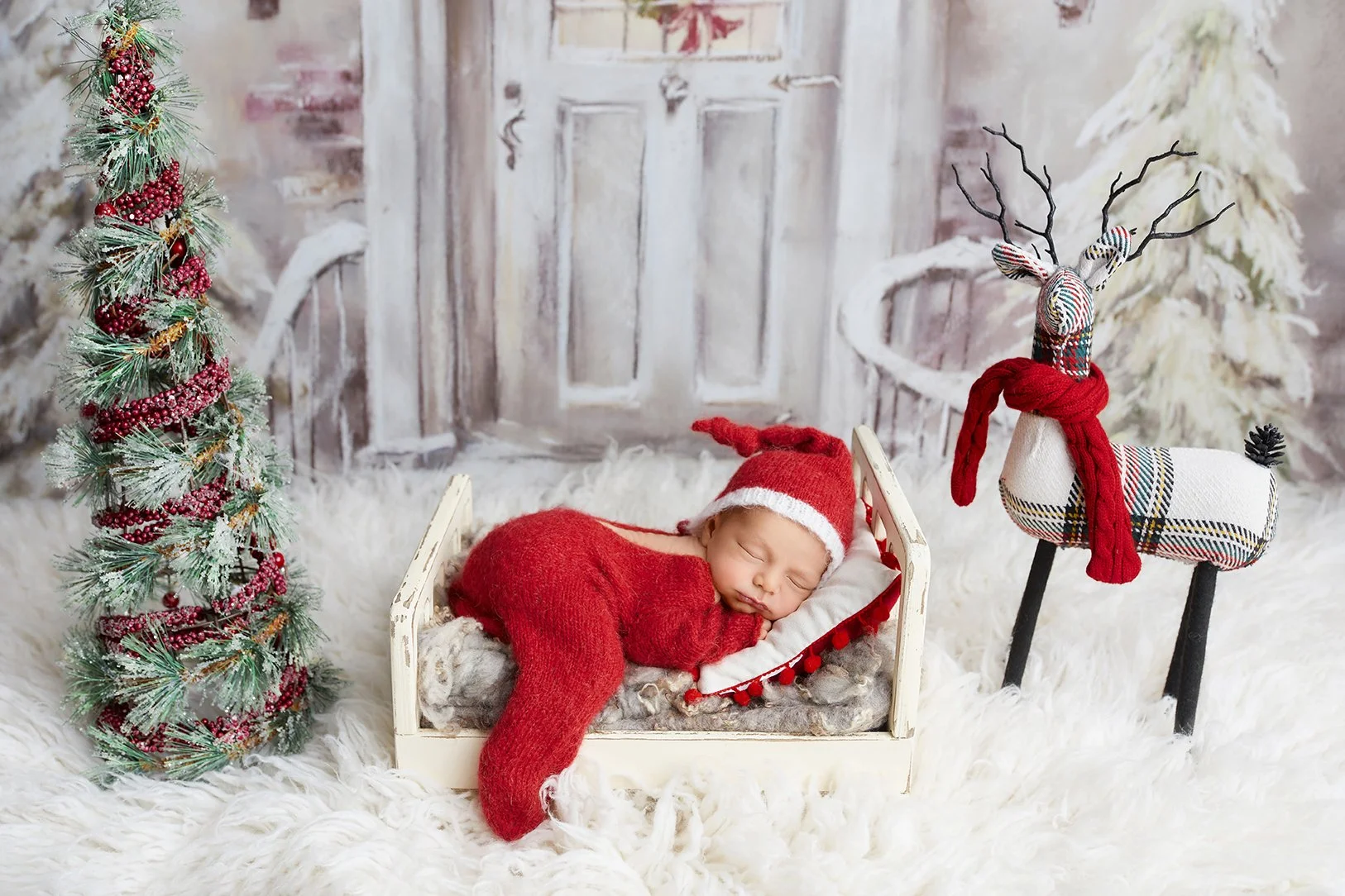 A baby wearing a red Christmas hat and outfit peacefully sleeping in a small wooden bed, surrounded by holiday decorations including a Christmas tree, a plaid reindeer plush, and a festive background with snow and winter elements.