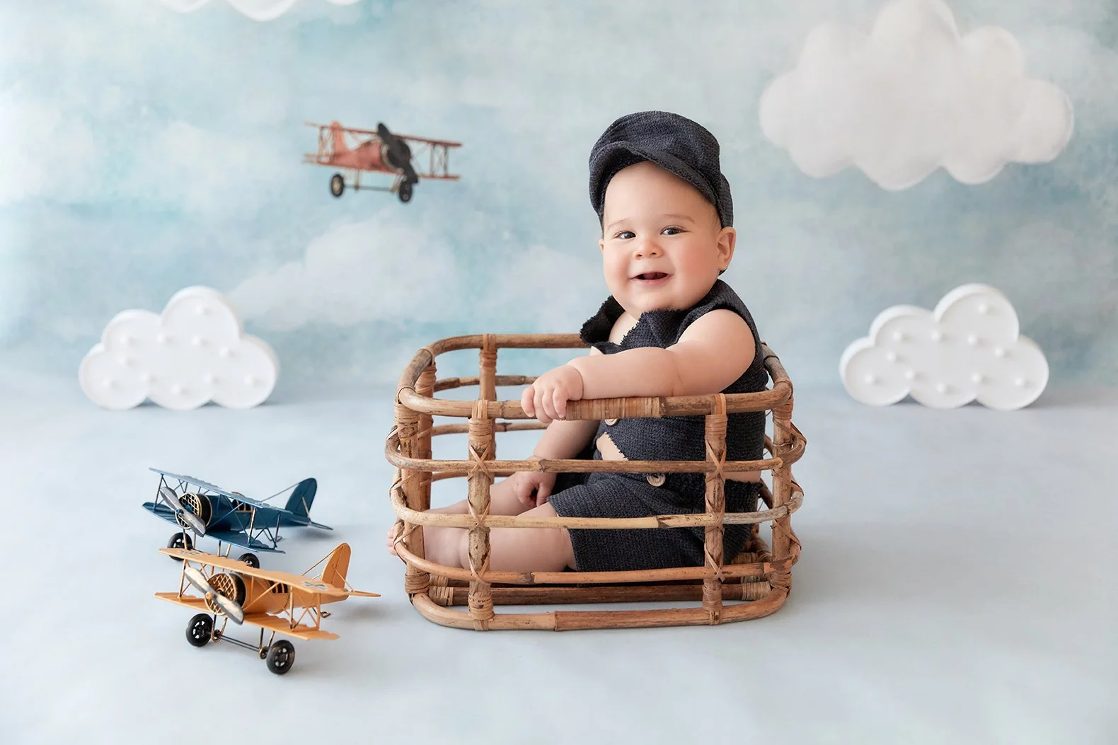 A smiling baby sitting inside a woven basket on a light blue surface, surrounded by toy airplanes and paper clouds in a sky background.