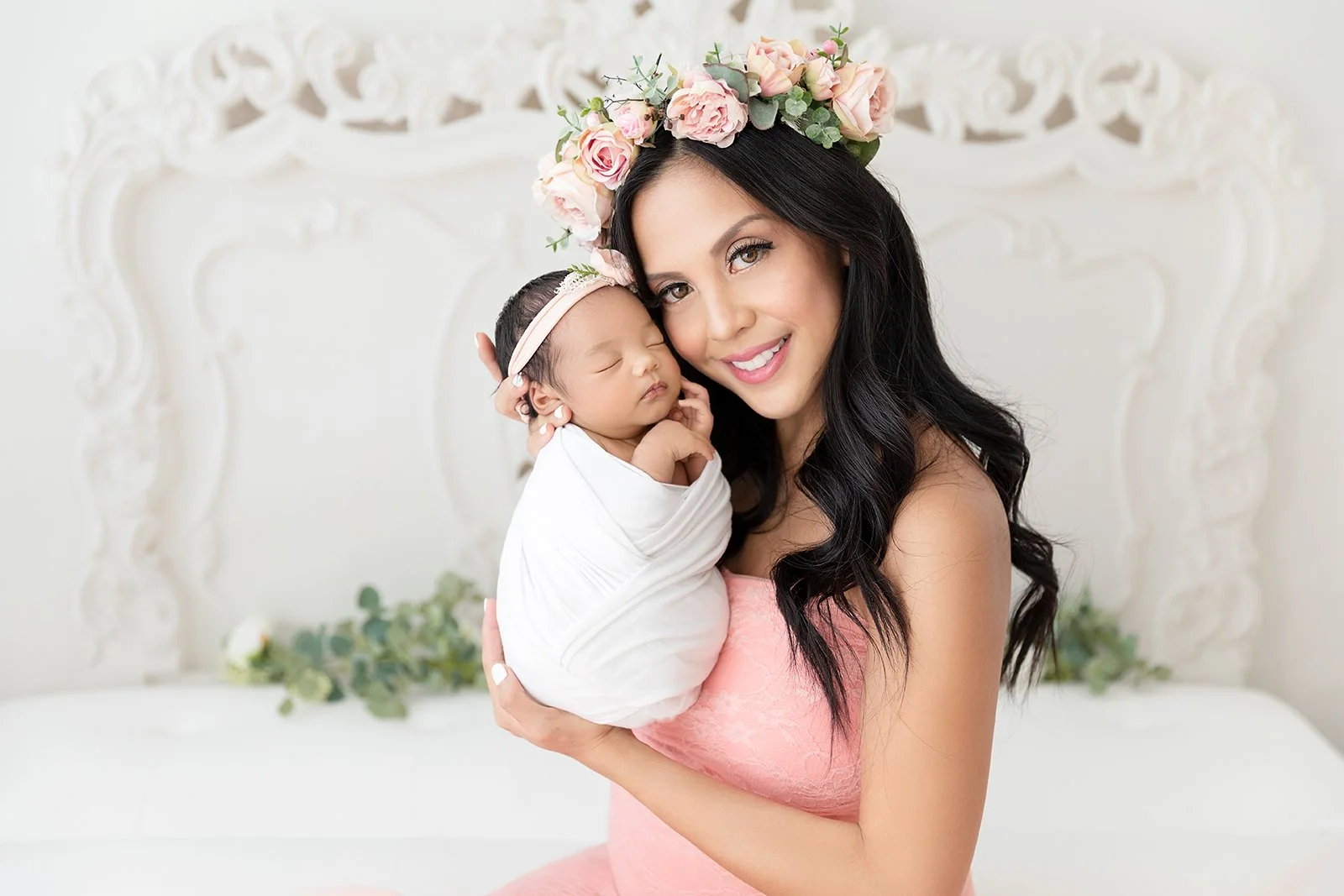 A smiling woman with long black hair wearing a pink dress and a flower crown holding a sleeping newborn baby wrapped in a white blanket.