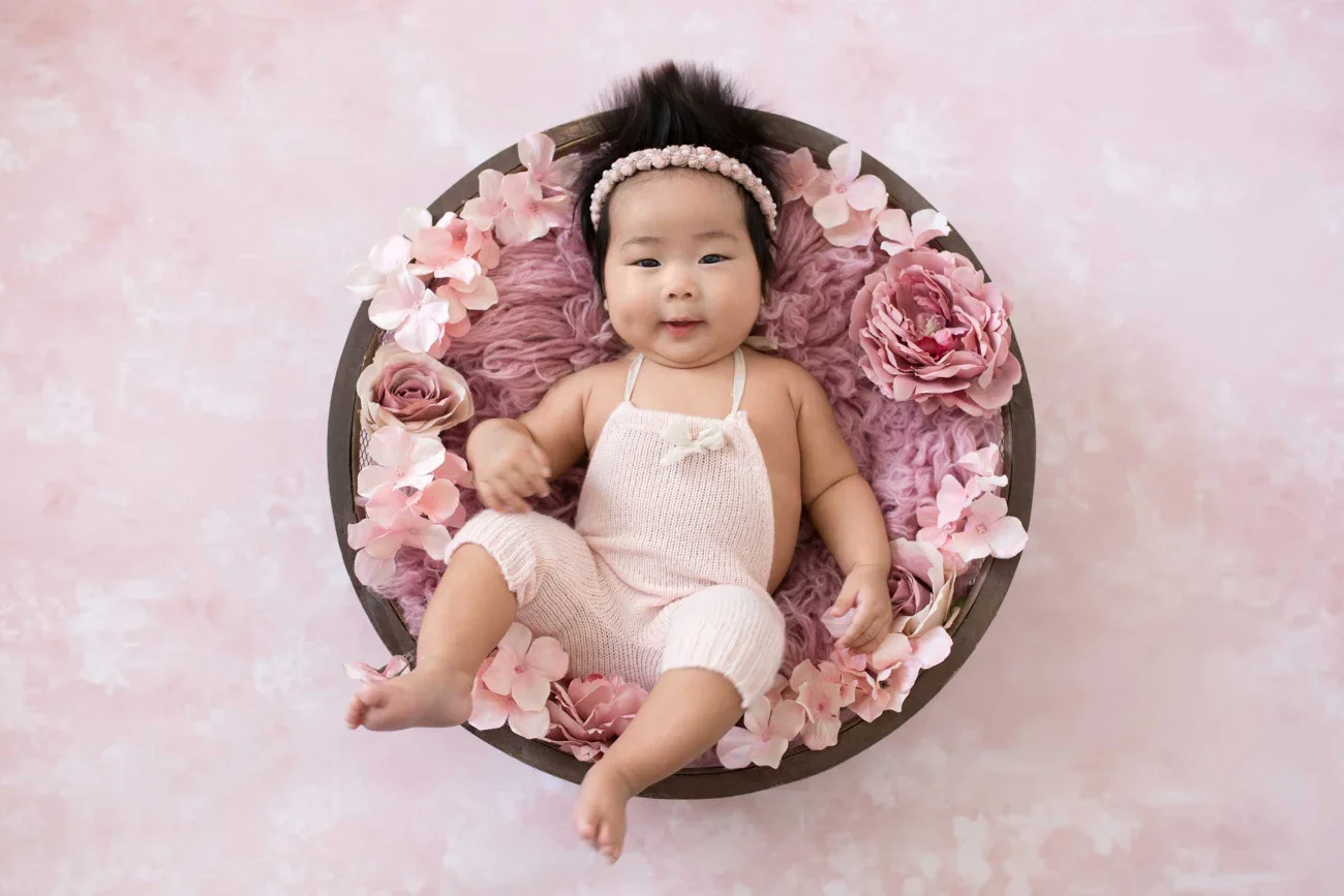 A smiling baby girl lying in a round basket filled with pink and purple flowers on a pink background.