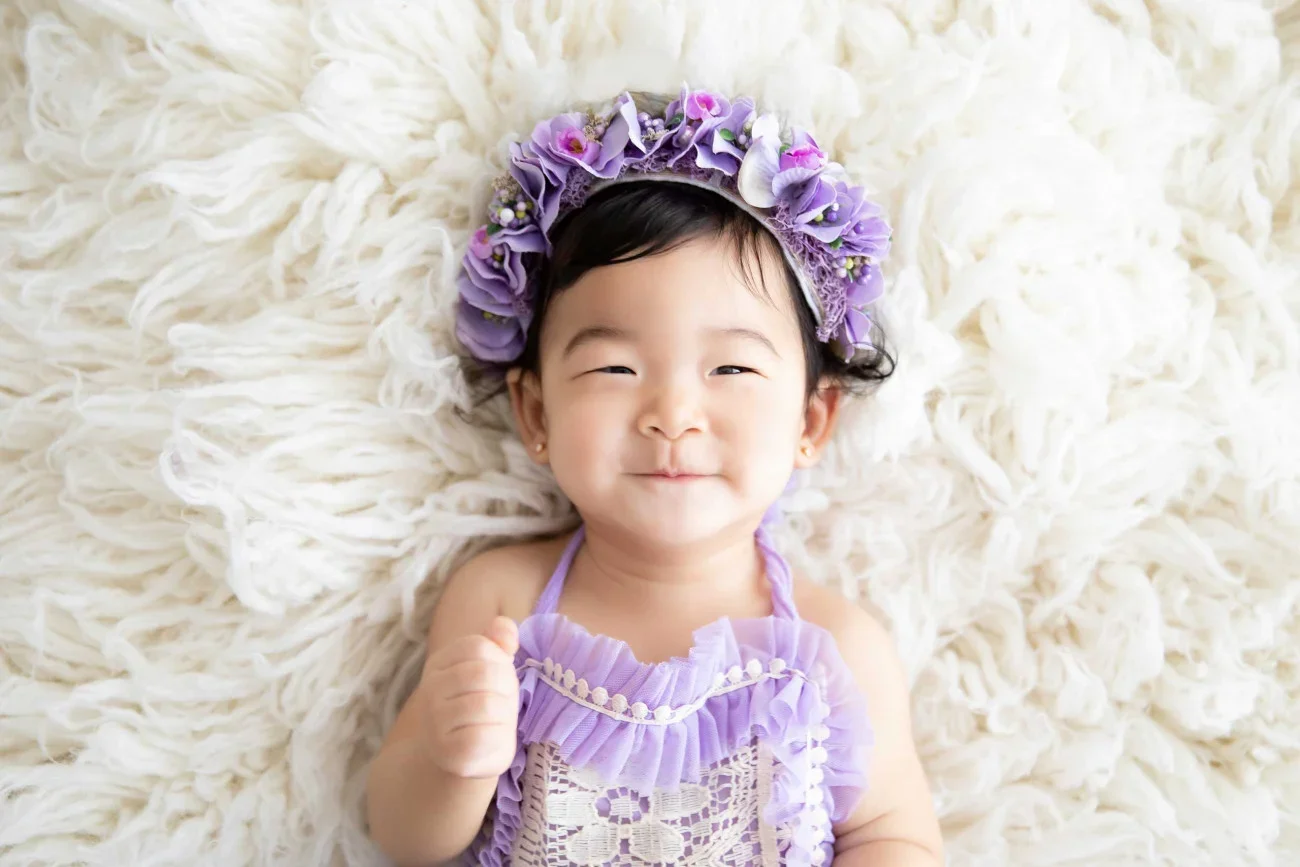 A smiling young girl lying on a white fluffy surface, wearing a purple ruffled dress and a purple flower crown.