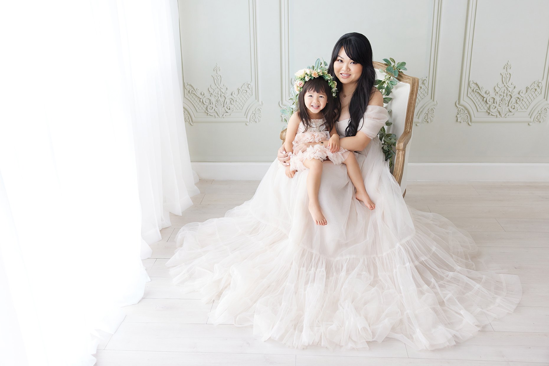 A woman in a wedding gown sitting on a chair with a young girl wearing a floral crown, both smiling and posing for the photo in a bright, elegant room with white decor.