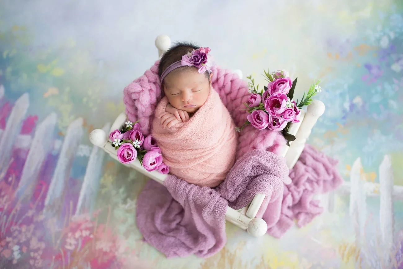 A newborn baby wrapped in pink cloth, sleeping on a pink blanket in a white cradle decorated with pink and purple flowers, on a colorful pastel background.
