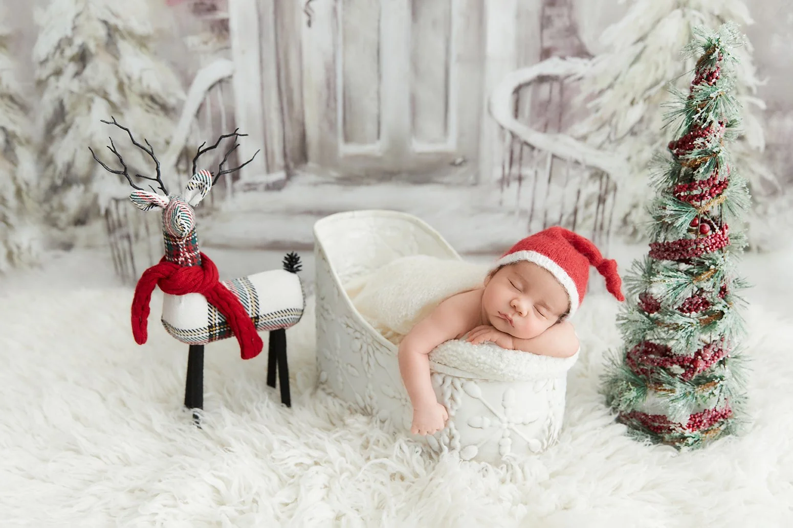 A sleeping baby in a Christmas scene, wearing a red Santa hat, lying on a white blanket, with a small fabric reindeer and Christmas tree decorations nearby.