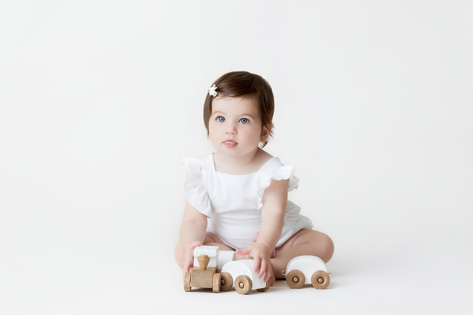 A young girl with brown hair, wearing a white dress, sitting on a white background playing with wooden toy cars and a train.