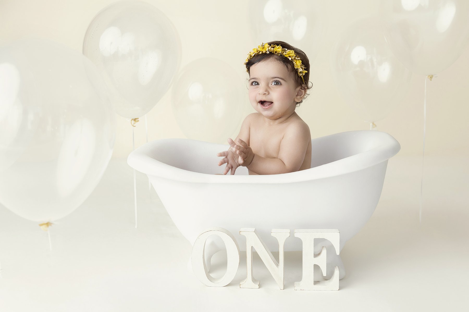 A smiling baby with a yellow flower headband sitting in a white bathtub surrounded by white balloons behind, with the word 'ONE' in large white letters in front of the bathtub.