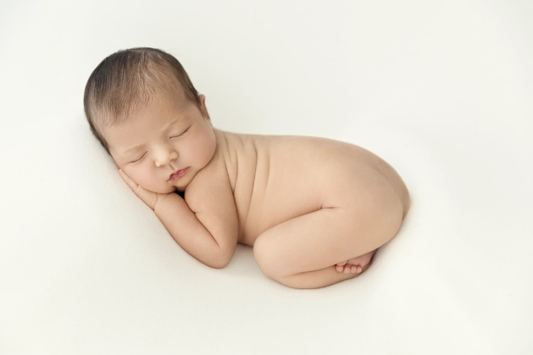 A sleeping newborn baby with closed eyes, resting on a white surface, curled up with its hands near its face.