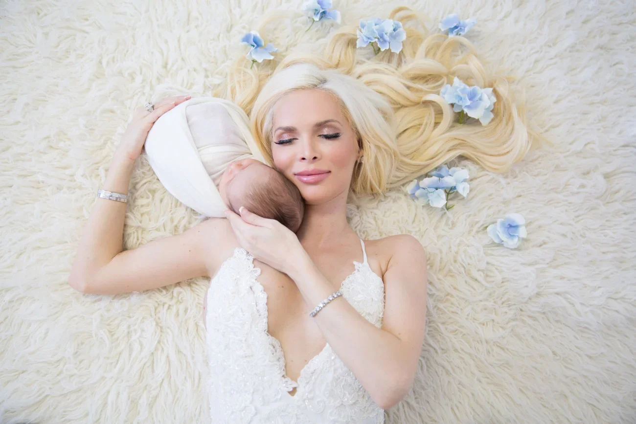 A blonde woman in a white dress lying on a fluffy cream-colored rug, holding a newborn baby with a hat and swaddling blanket, surrounded by light blue flowers.