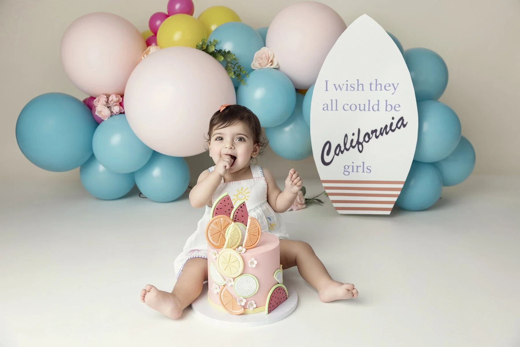A young girl sitting on the floor with a pink fruit-themed cake in front of her, surrounded by balloons and a sign that says, 'I wish they all could be California girls'.