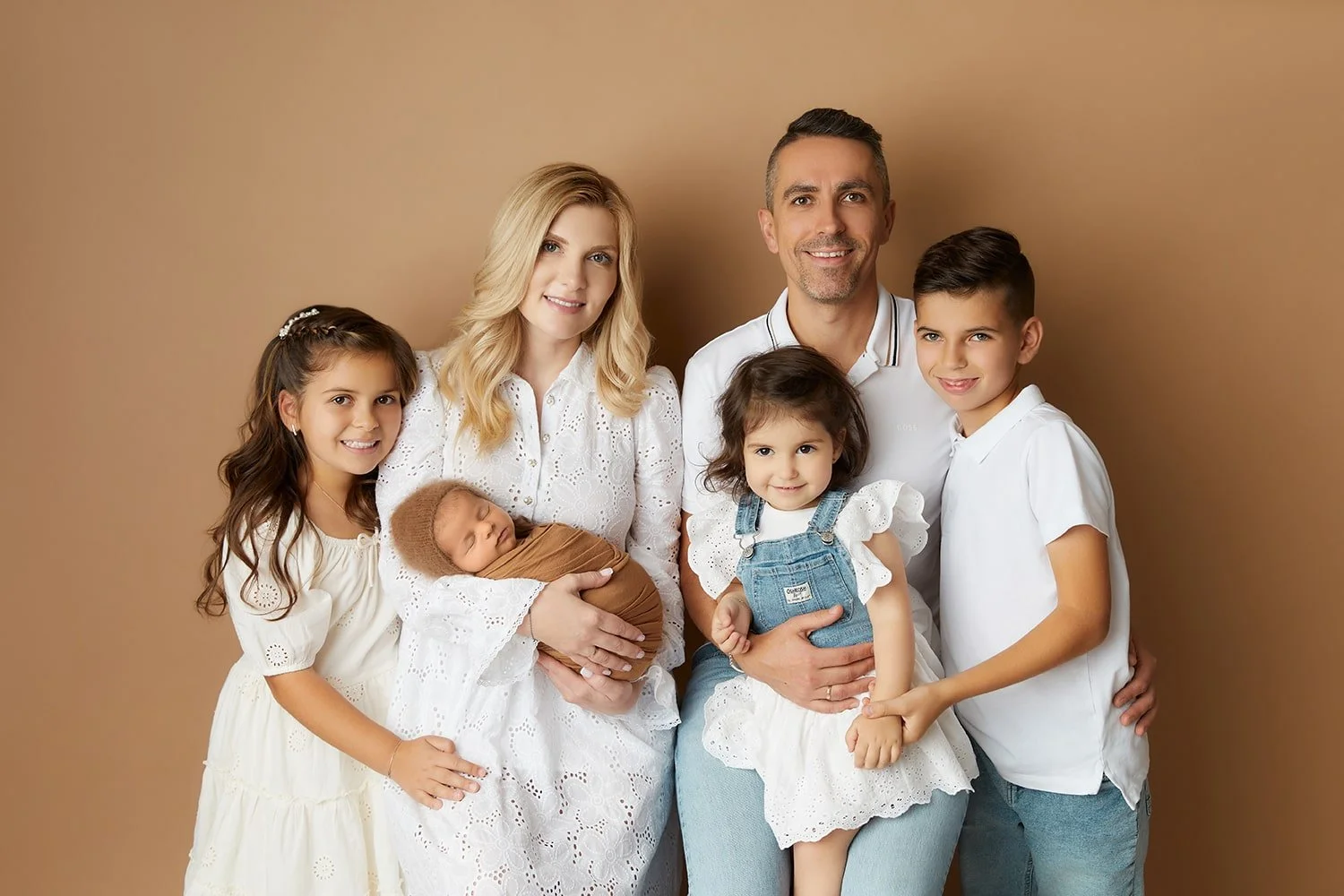 A family of seven posing together in front of a beige background. The group includes a woman, a man, four girls, and a newborn baby. They are all smiling, dressed in white and light-colored clothing.