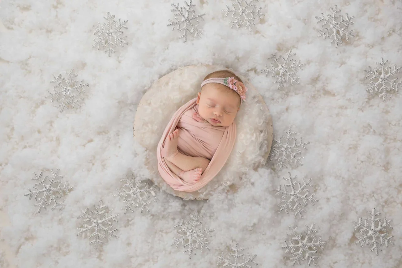 A baby girl is sleeping on a soft white surface decorated with silver snowflakes, wrapped in a light pink swaddle, wearing a pink floral headband.