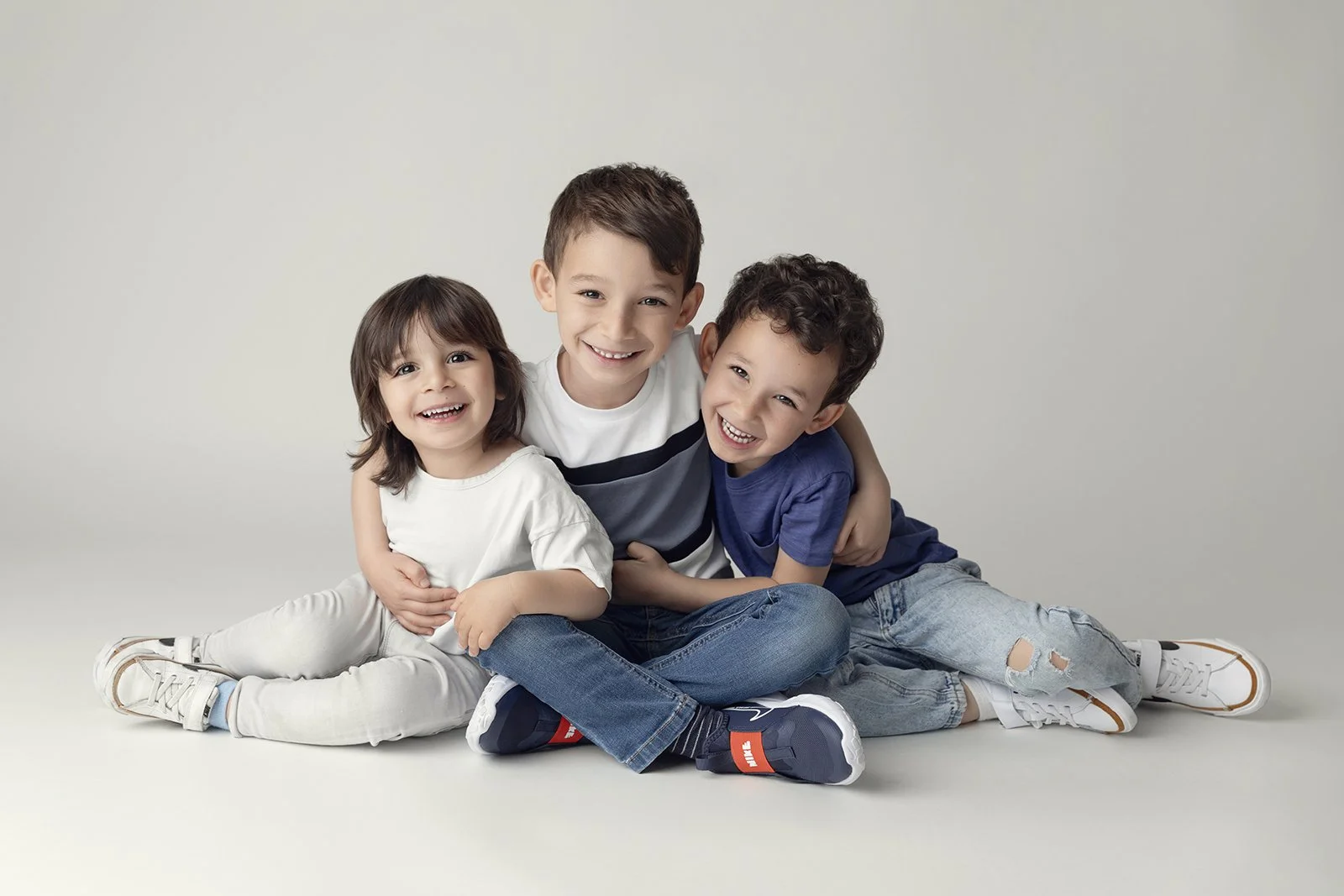 Three children sitting on the floor, hugging each other and smiling, against a plain light background.