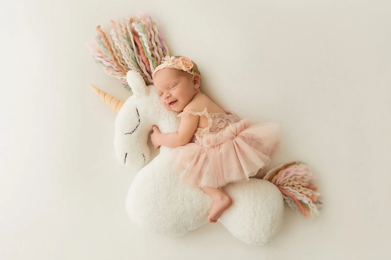 A baby girl wearing a pink lace dress and tulle skirt, lying on a plush white unicorn stuffed animal with a multicolored yarn mane and tail, against a plain light background.