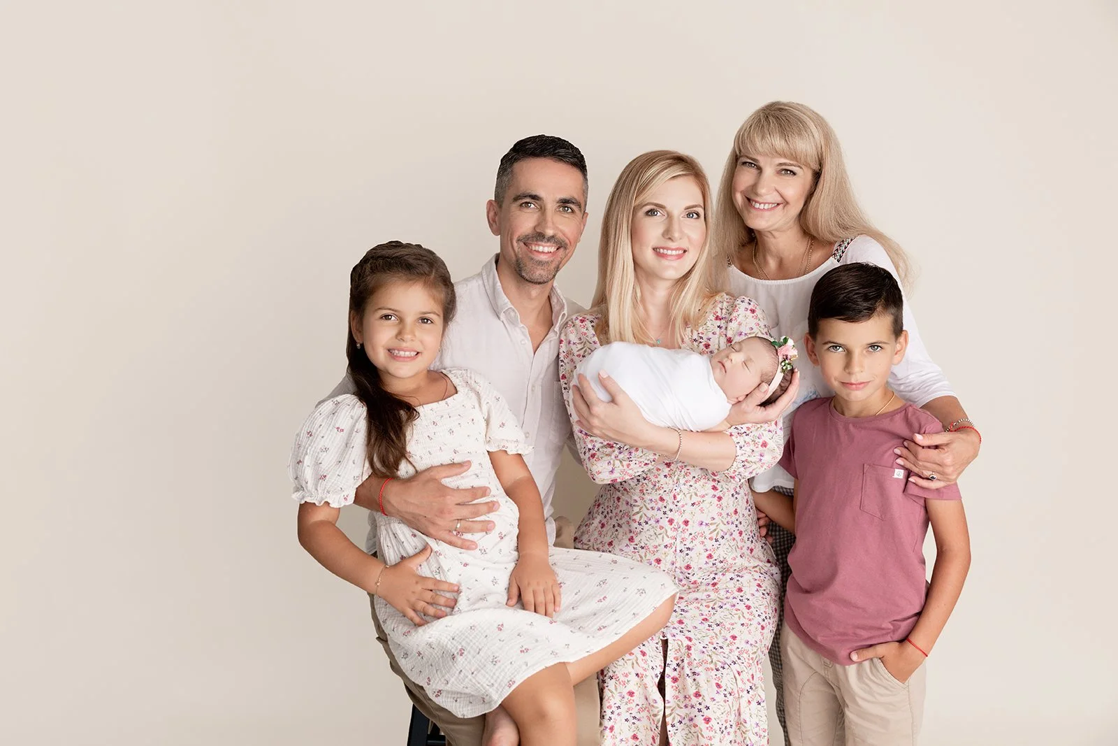 A family portrait of six people, including two young children, a mother holding a newborn, a father, and two older women, all smiling against a plain light background.