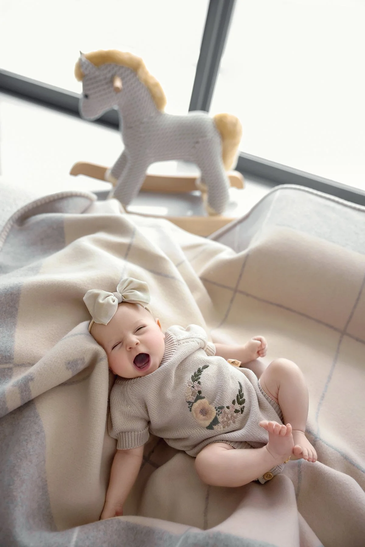 A baby girl lying on a blanket, yawning or laughing, wearing a beige dress with floral embroidery and a large bow headband. In the background, a stuffed unicorn toy is placed on a wooden hanger near a window.