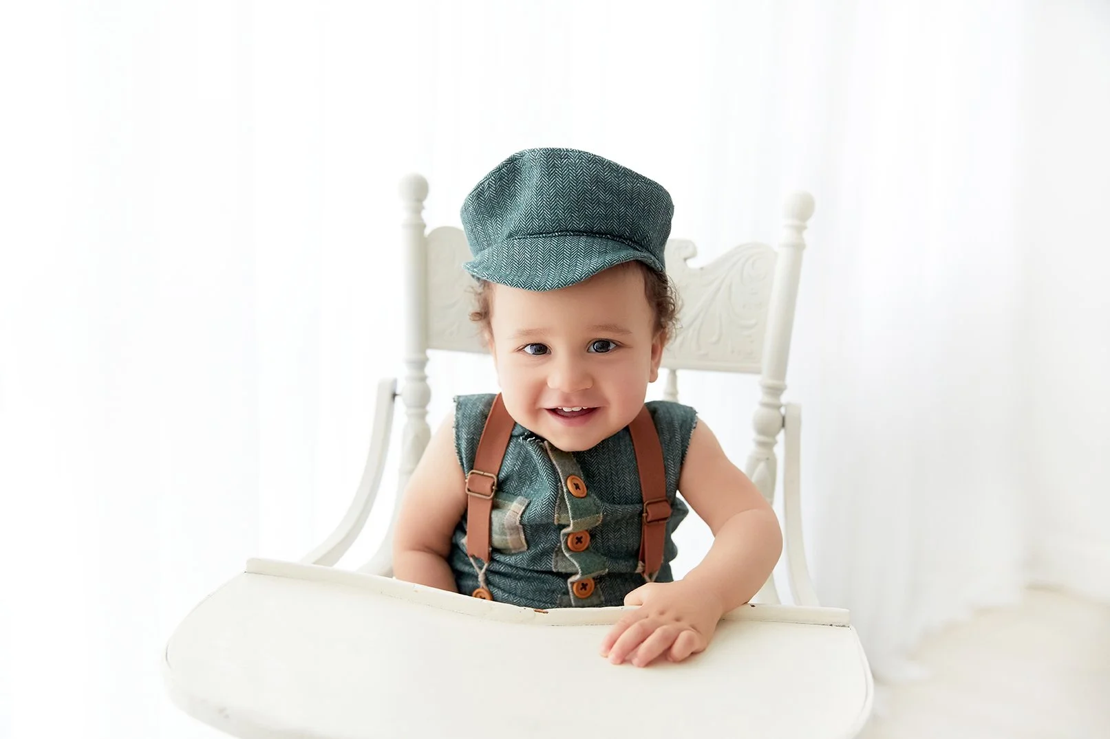 A smiling baby with curly hair, wearing a matching denim hat and vest with brown suspenders, sitting in a white high chair against a bright white background.