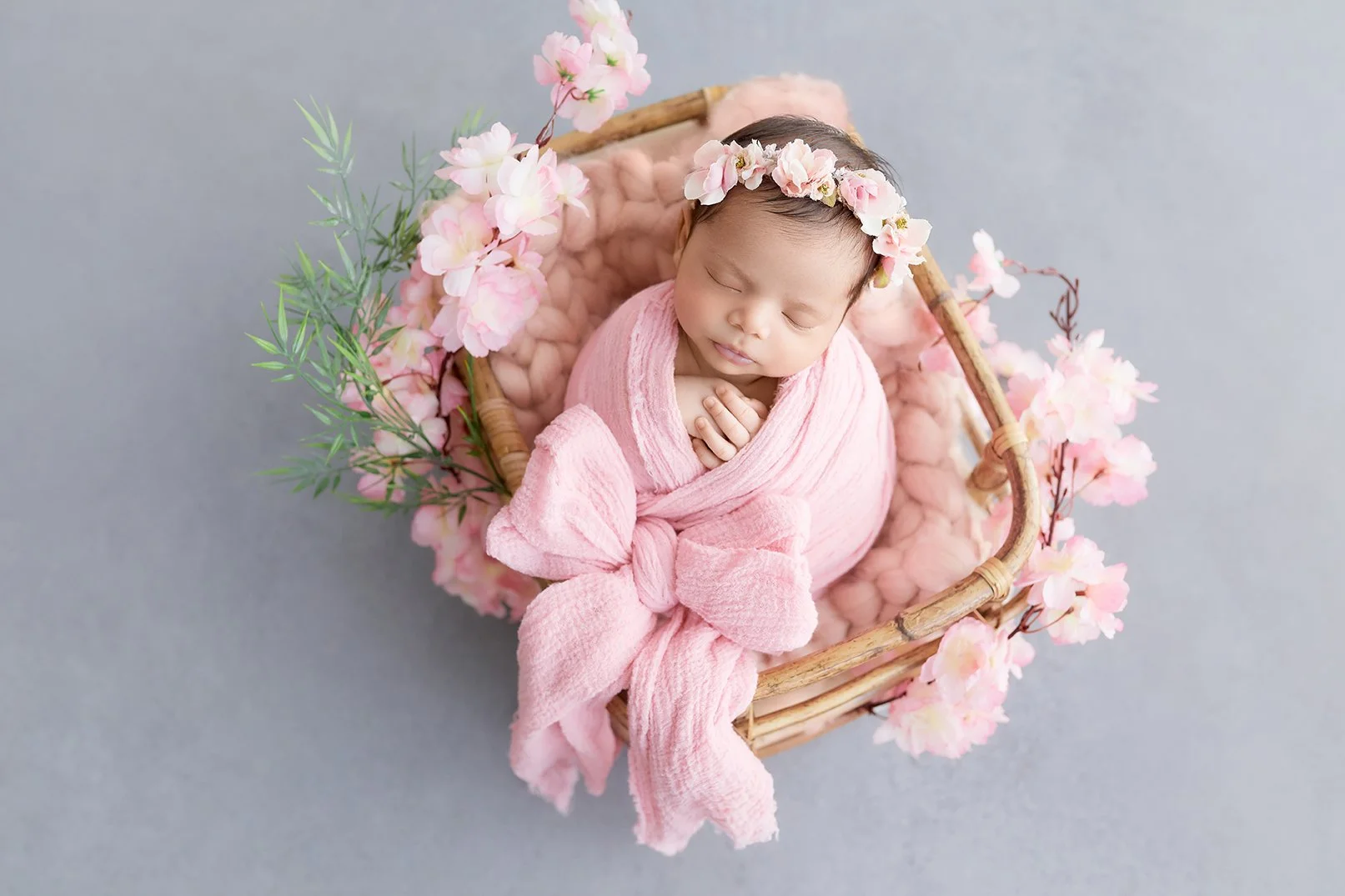 A sleeping baby girl wrapped in a pink blanket and wearing a pink floral headband, lying in a wicker basket decorated with pink flowers and green leaves on a soft gray background.
