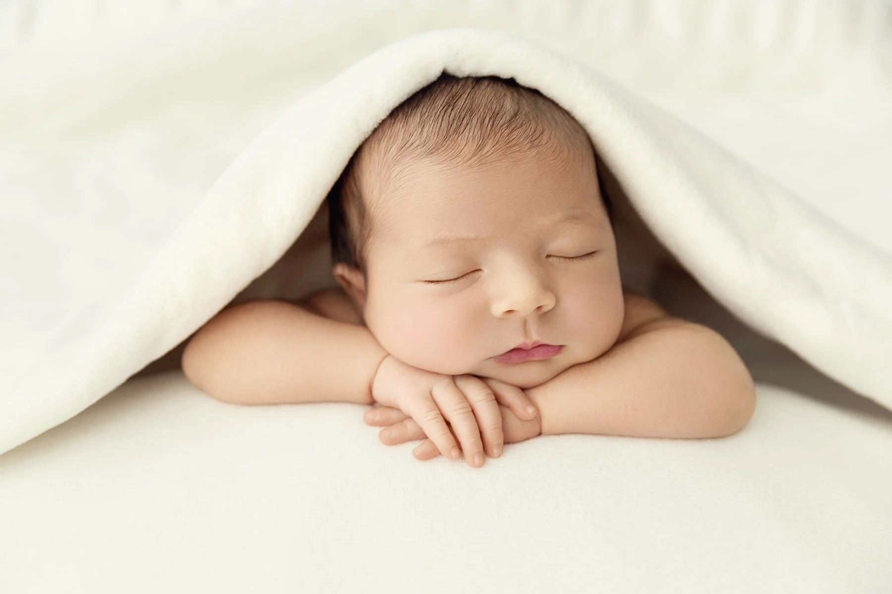 A sleeping baby with arms crossed under chin, covered partially by a white blanket