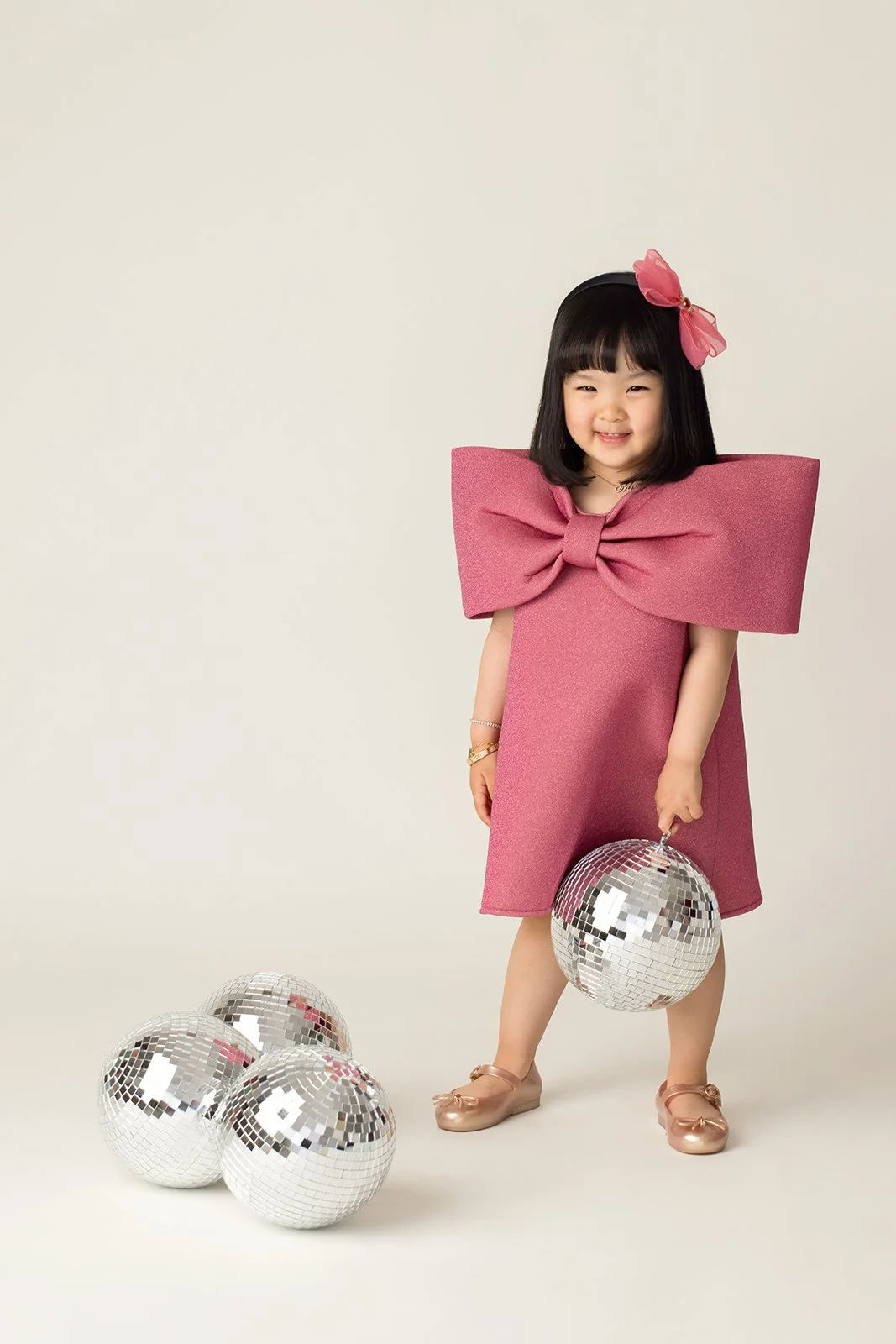 Young girl in a pink dress with a large bow, holding a disco ball, surrounded by three additional disco balls on the floor, smiling at the camera.
