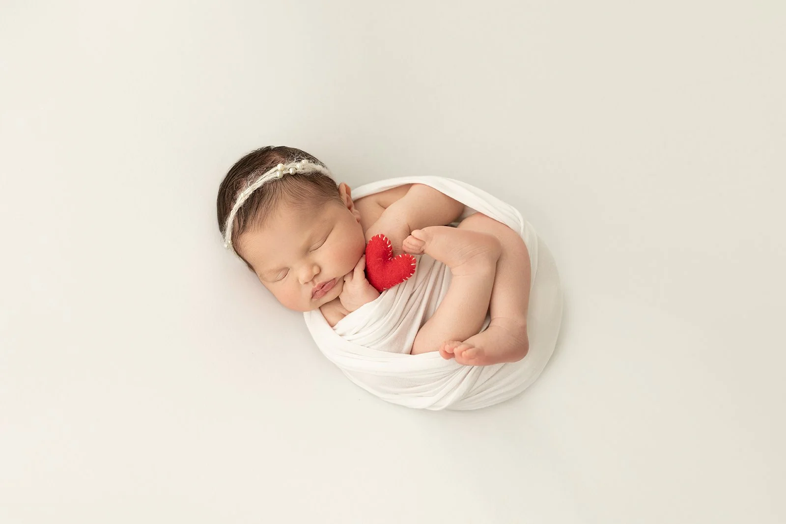 Newborn baby sleeping peacefully, wrapped in white fabric, holding a small red heart plush toy, with a white headband, on a plain light background.