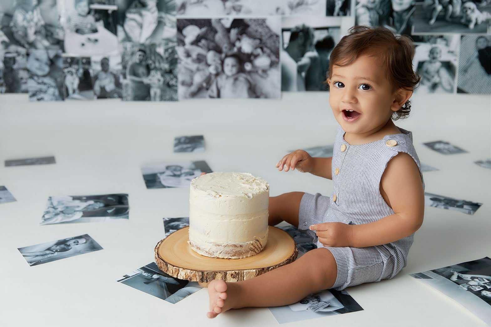 A young child with dark hair sitting on a white surface, celebrating a birthday with a small white frosted cake on a wooden slab. The backdrop features black-and-white photos pinned to a wall. The child's expression is joyful and surprised.