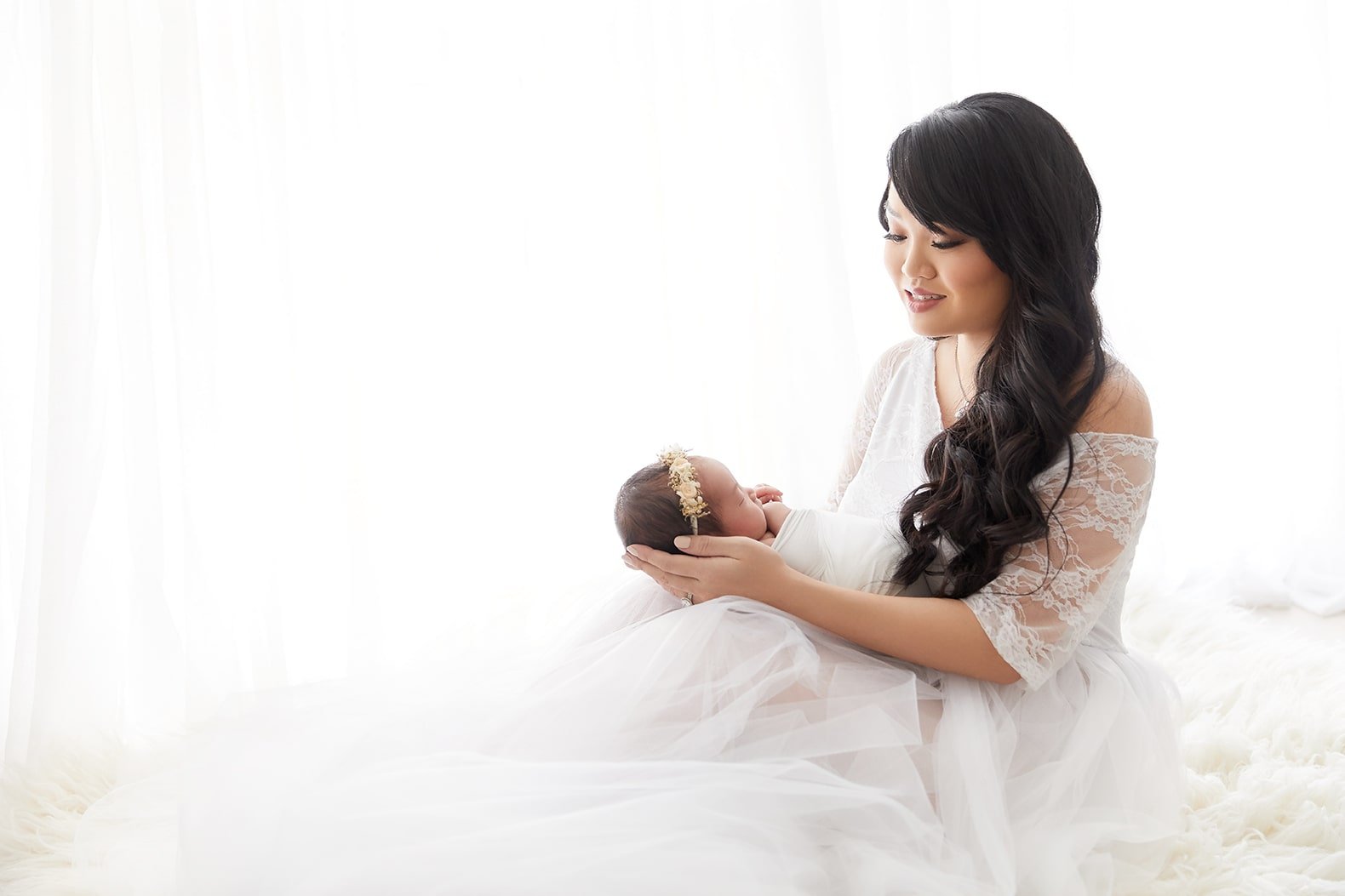 A woman with long dark hair in a white lace dress holds a sleeping baby dressed in white with a floral headband, both on a fluffy white surface with a bright background.