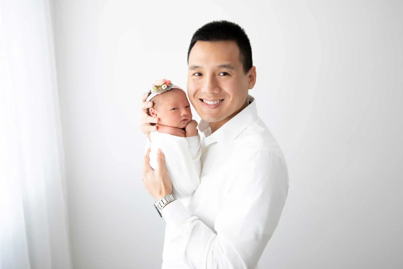 A smiling man in a white shirt holding a newborn baby girl with a floral headband against a plain white background.
