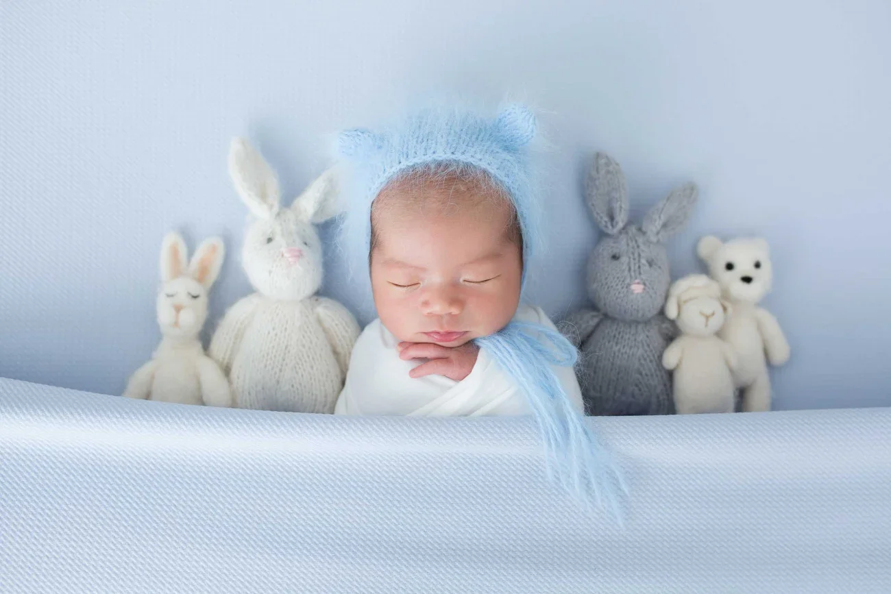 A sleeping baby wearing a light blue knit hat, surrounded by knitted bunny and bear stuffed animals.
