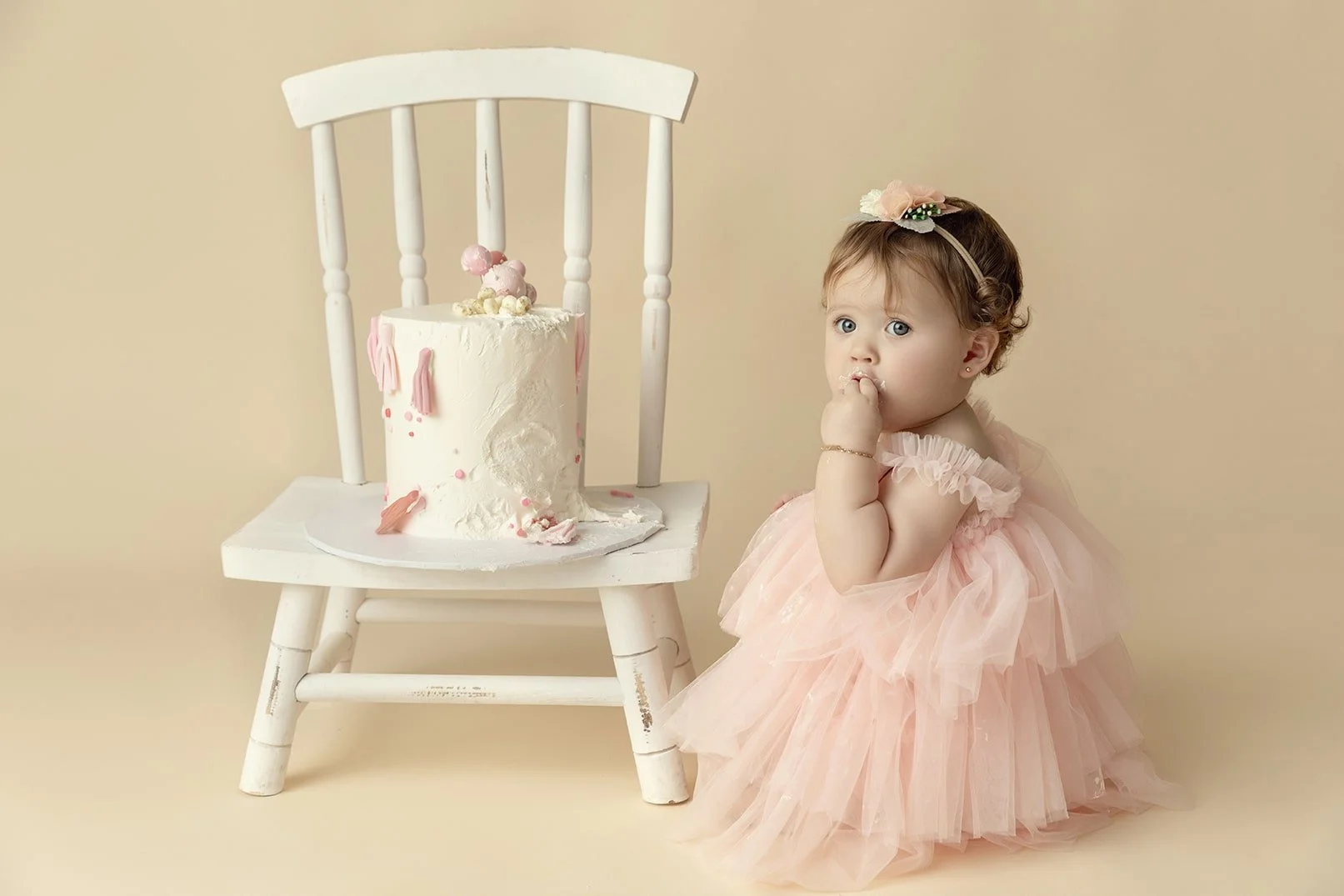 A young girl in a pink tutu dress sitting on the floor next to a white wooden chair and a birthday cake with pink decorations.