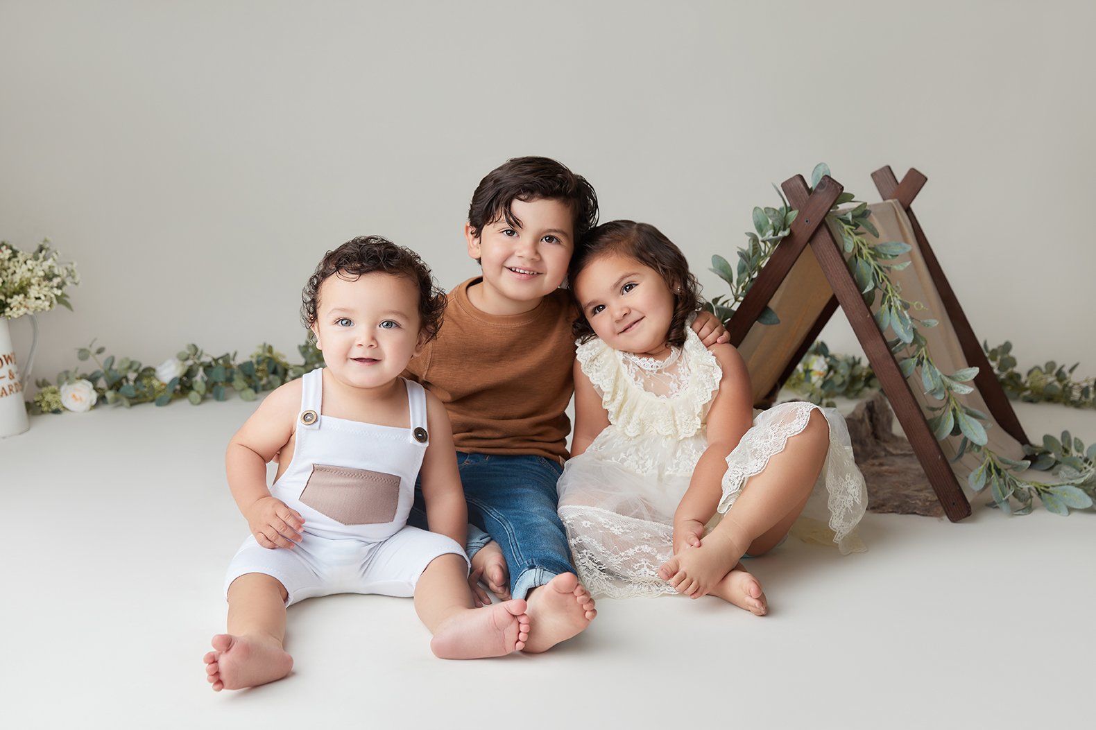 Three children sitting on the floor with a decorative background, including a small wooden ladder, greenery, and flowers.