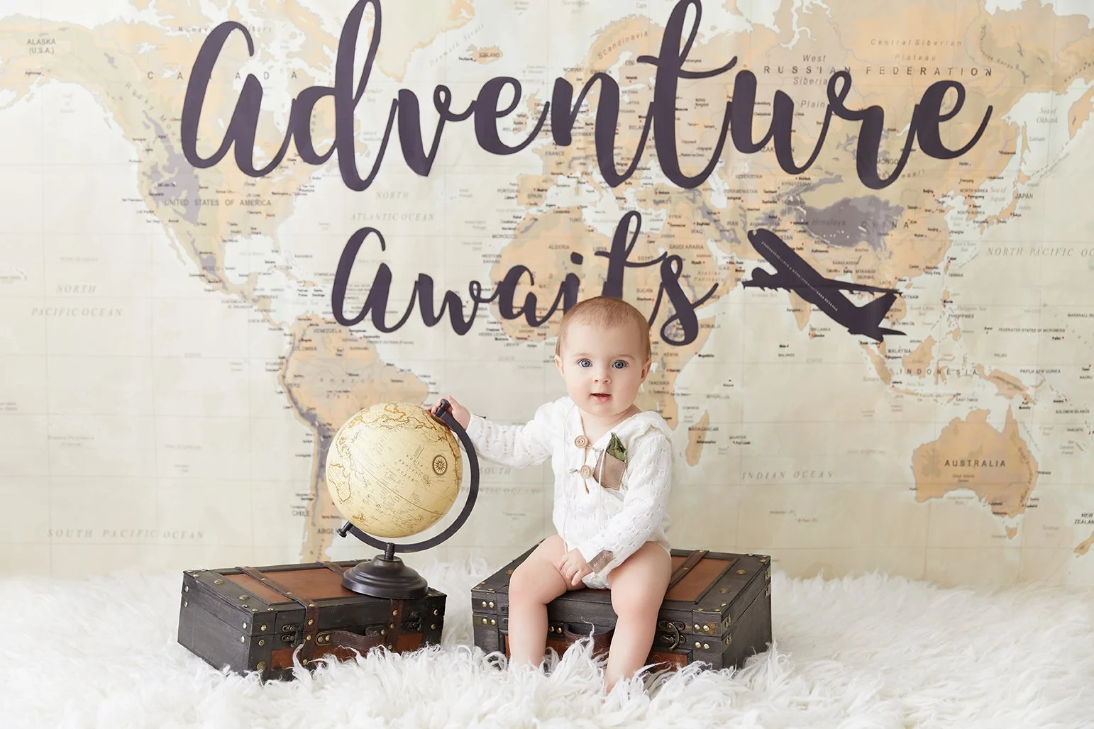 A baby sitting on a trunk in front of a world map backdrop with the text 'Adventure Awaits' and an airplane graphic. The baby holds a globe and sits on a white furry rug.