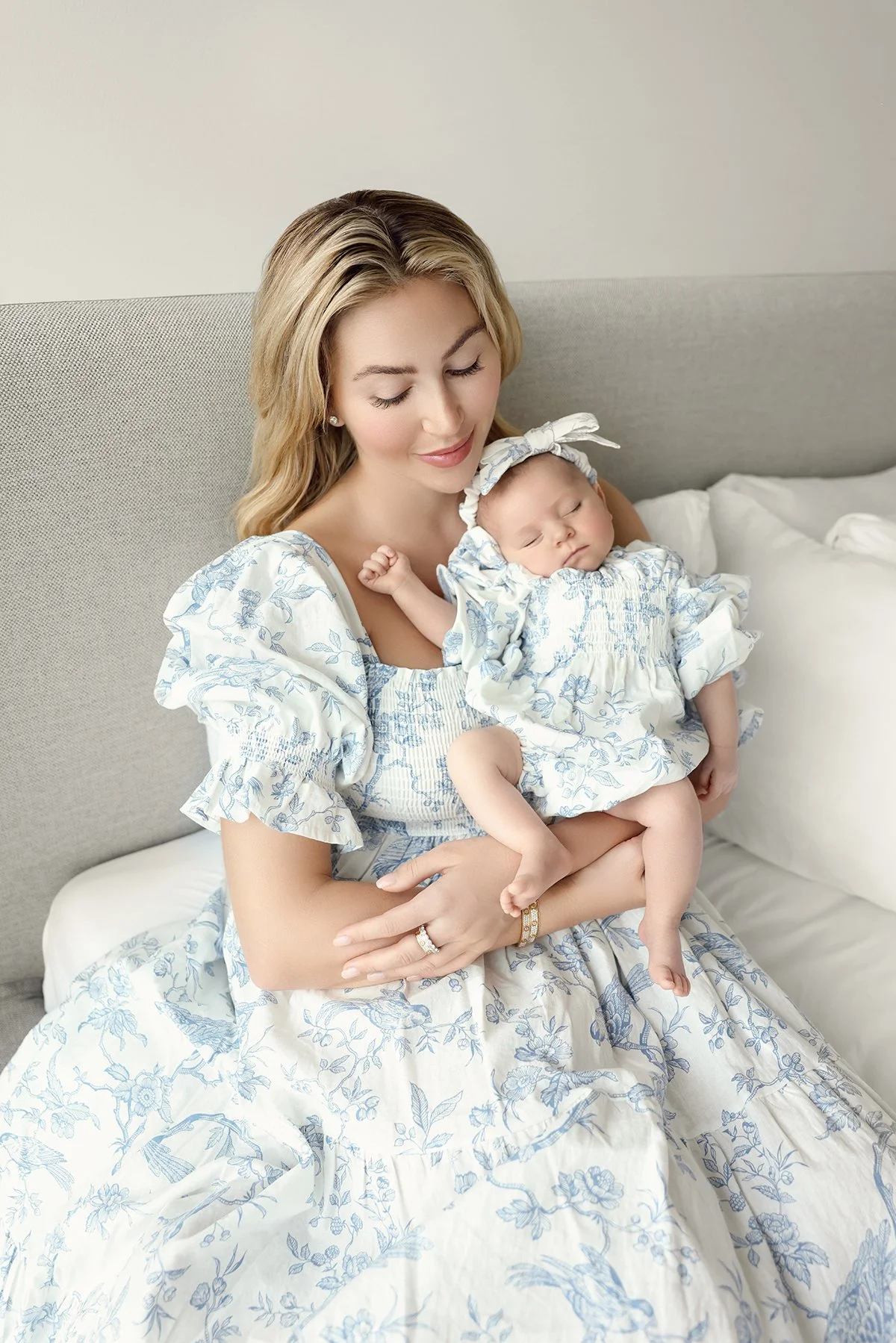 A woman holding a sleeping baby girl on a bed, both dressed in matching white and blue floral outfits, in a light-colored room.