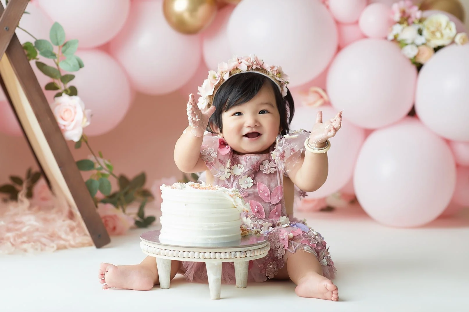 Smiling baby girl celebrating her first birthday sitting in front of a cake, wearing a floral dress and headband, with pink and white balloons and flowers in the background.