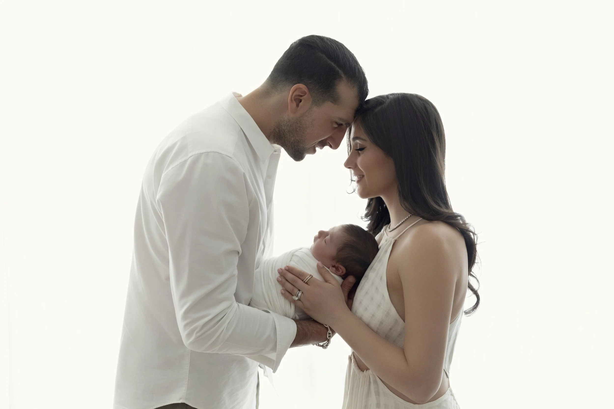 A couple holding their newborn baby, touching foreheads, in a bright, softly lit room.